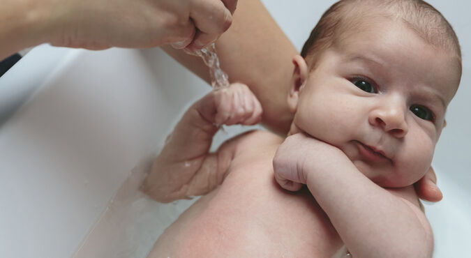Bebê tomando banho na banheira com um adulto segurando-o por trás do pescoço