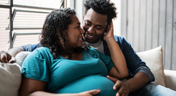 Mulher grávida junto com parceiro sentados em sofá em momento de carinho