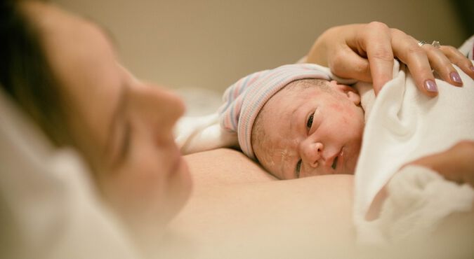 Imagem de gestante deitada com seu bebê vestindo touca e com manta nos braços logo após o parto
