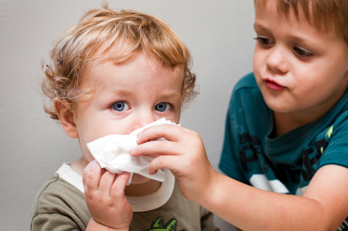 Criança branca com congestão nasal tendo nariz assoado por  menino maior.